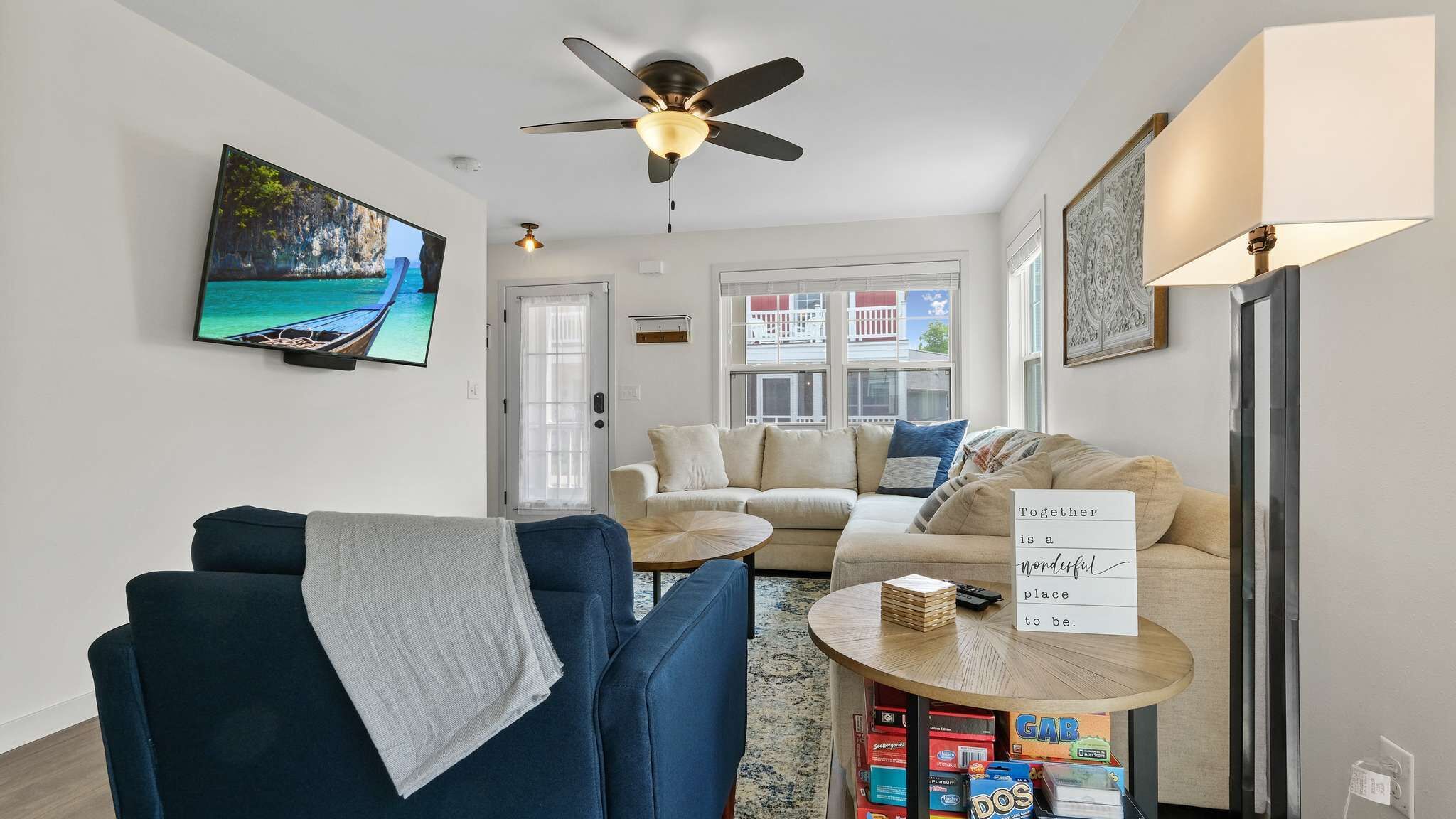 A living room with a sofa, armchair, TV, ceiling fan, and a table with a sign.