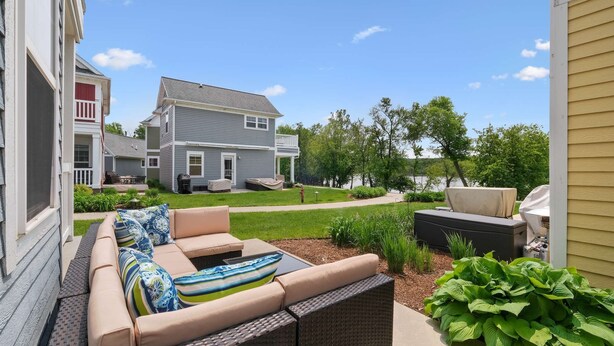 Eine Terrasse mit Gartenmöbeln, einem überdachten Grill und Blick auf ein Haus und einen See.