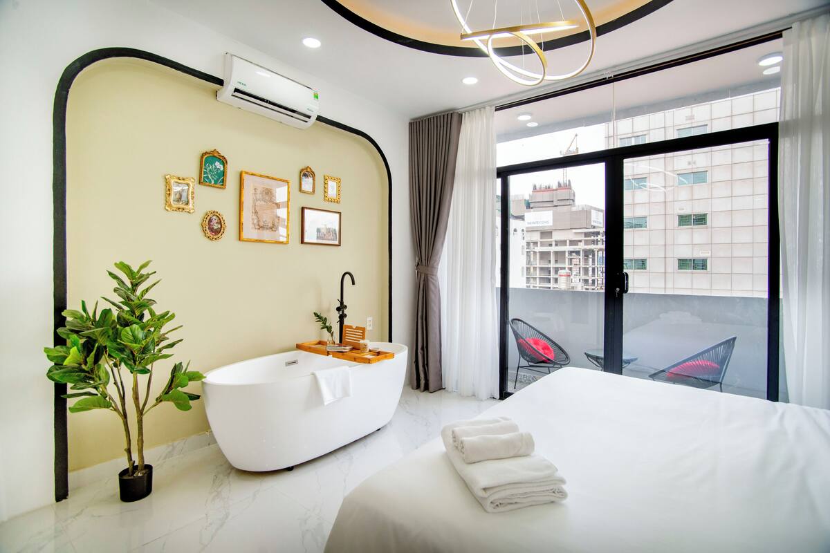 Ein modernes Hotelzimmer mit einem großen Bett, einer freistehenden Badewanne, einem Blumentopf und Blick auf die Stadt durch große Fenster.