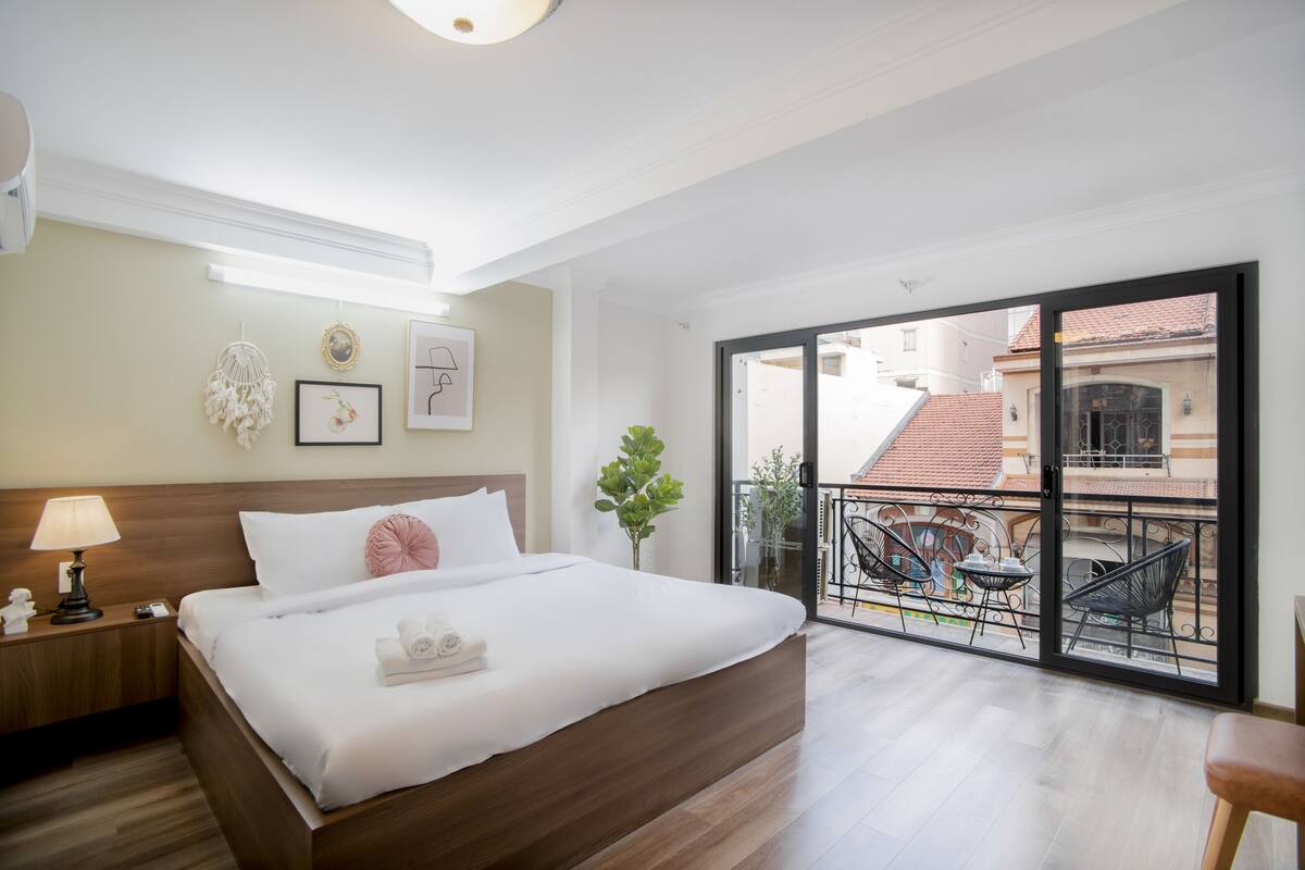 Ein Hotelzimmer mit einem großen Bett, einem Nachttisch mit Lampe, einem Balkon mit Tisch und Stühlen und Blick auf Gebäude.