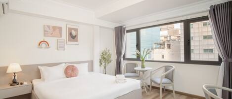 A hotel room with a large bed, a small table with chairs, a window with curtains, and artwork on the wall.