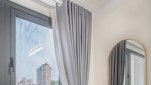 Een kamer met raam, grijze gordijnen, een spiegel en uitzicht op gebouwen.