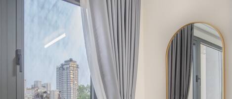 Une pièce avec une fenêtre, des rideaux gris, un miroir et une vue sur des immeubles.