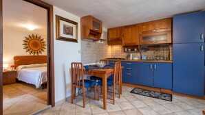 A compact kitchen with wooden cabinets, a dining table with chairs, a bed in the adjacent room, and a sun decoration on the wall. - Holiday Home 'Casetta Brandinu' with Mountain View, Balcony & Wi-Fi (Sassari)