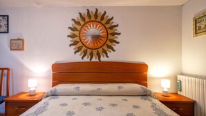 A bedroom with a wooden headboard, a floral bedspread, two bedside lamps, and a wall-mounted sun decoration. - Holiday Home 'Casetta Brandinu' with Mountain View, Balcony & Wi-Fi (Sassari)