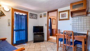 A room with a bed, a TV, a dining table with chairs, a kitchen area, and a bathroom visible through an open door. - Holiday Home 'Casetta Brandinu' with Mountain View, Balcony & Wi-Fi (Sassari)