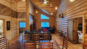 Interior de una cabaña de troncos con comedor, asientos y un ventanal grande.