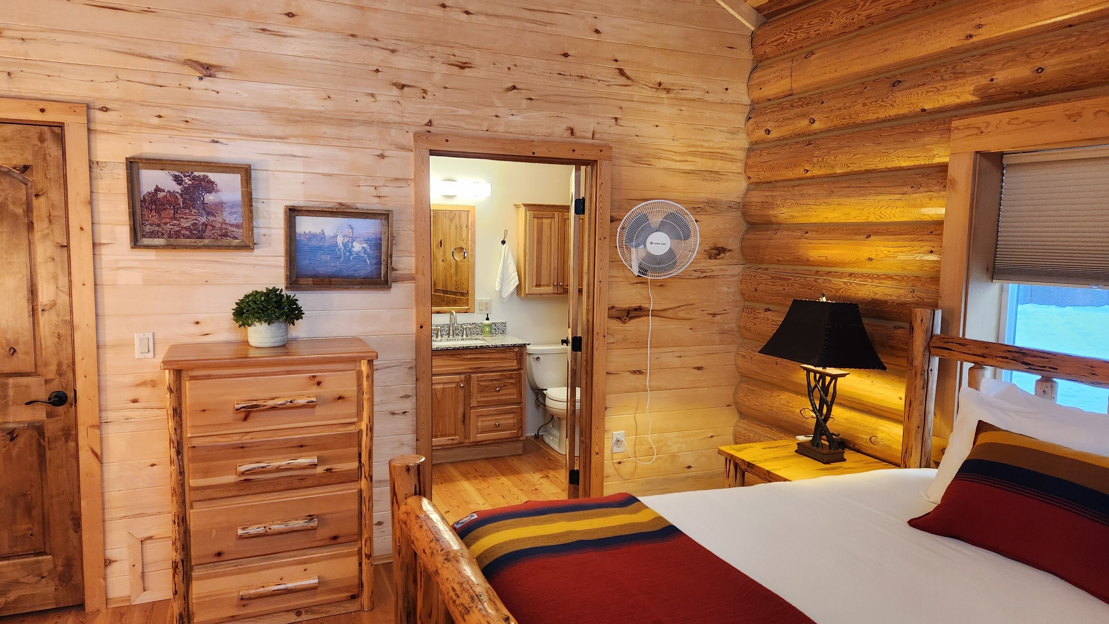 A wooden cabin interior with a bed, dresser, fan, and framed pictures on the wall.