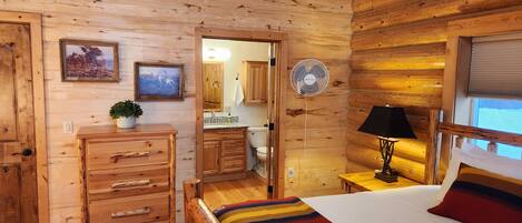 A wooden cabin interior with a bed, dresser, fan, and framed pictures on the wall.