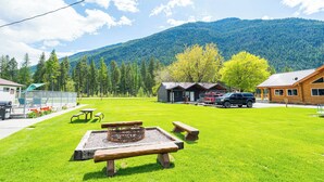 Un camping con mesas de picnic, una fogata, cabañas y montañas al fondo.