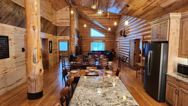 Interior de una cabaña de troncos con comedor, cocina y un amplio ventanal.