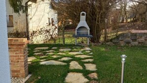 A stone pathway leading to a barbecue area with a chimney, surrounded by a grassy yard and a brick wall. - Apartment with panoramic lake view, sauna, close to hiking trails. (Orfu)