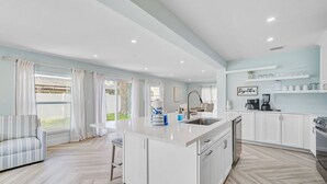 A modern kitchen with white cabinets, a central island, and a dining area with striped furniture. - Private Pool Retreat Near Anna Maria Island -Spa - Optional Pool Chiller-EV Charger (Bradenton)
