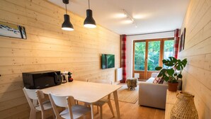 L'intérieur d'un chalet accueillant, avec une table à manger en bois, des chaises blanches, un canapé et une grande fenêtre donnant sur un paysage verdoyant.