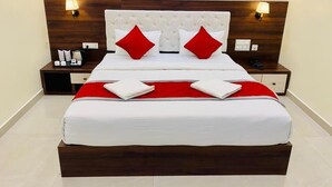 A neatly made bed with a wooden headboard, two red pillows, and a white bedspread. There are two bedside tables with lamps and a small tray of toiletries. - PPH Living Gnr Grand Luxury Rooms (Madanapalle)