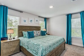 Ein Schlafzimmer mit einem Holzkopfteil, einem Bett mit gemusterten Betttextilien, einem Nachttisch mit Lampe und lindgrünen Vorhängen.