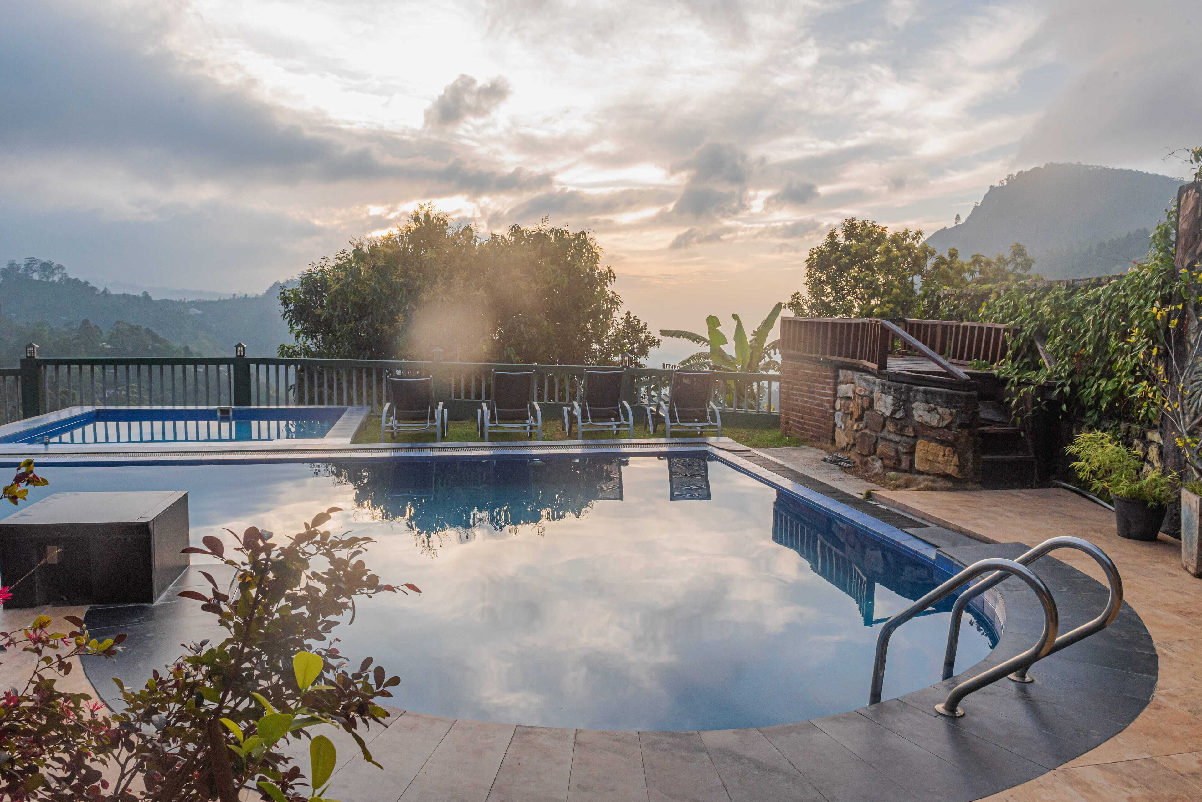 Piscina con pared de piedra y barandillas metálicas, rodeada de vegetación y vistas a colinas.