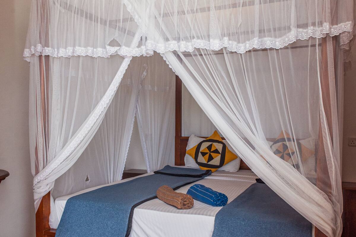 A bed with a wooden frame and white bedding, a blue blanket, and a decorative pillow with a geometric pattern.