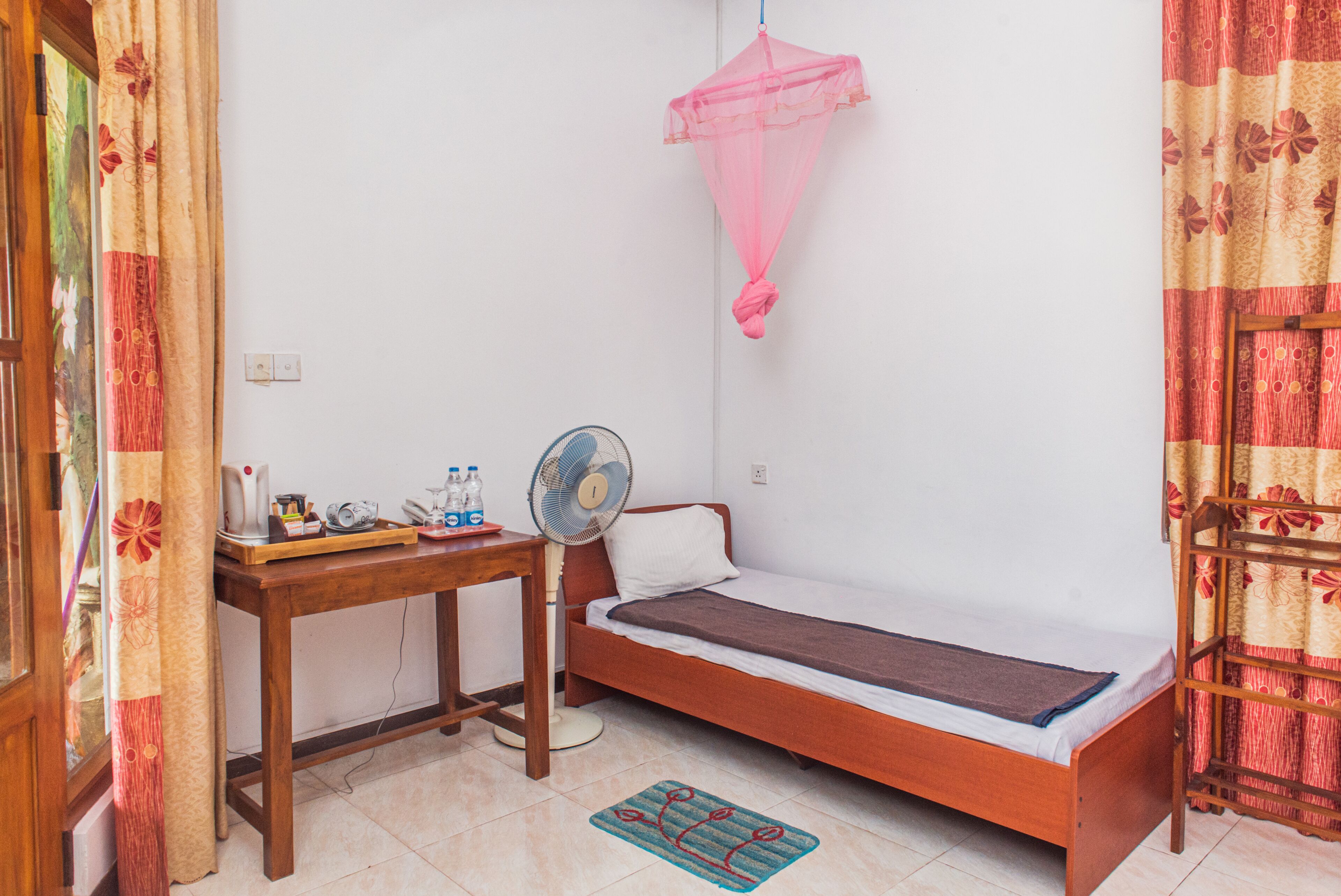 Ein kleines Zimmer mit einem Einzelbett, einem Holzbett, einem Tisch mit einem Ventilator, einem Wasserspender und einer Steckdose.