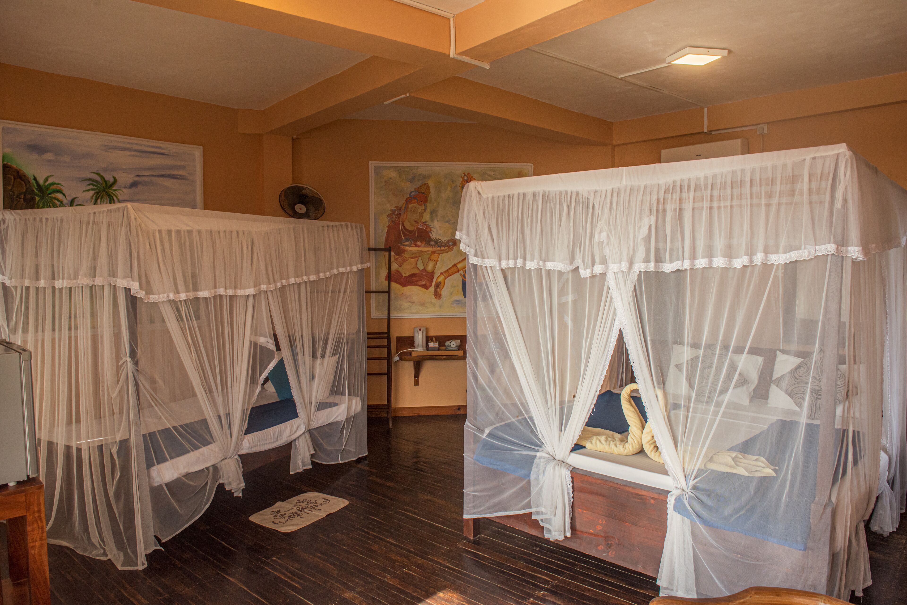 Two single beds with white canopies, wooden flooring, a painting on the wall, and a ceiling fan.