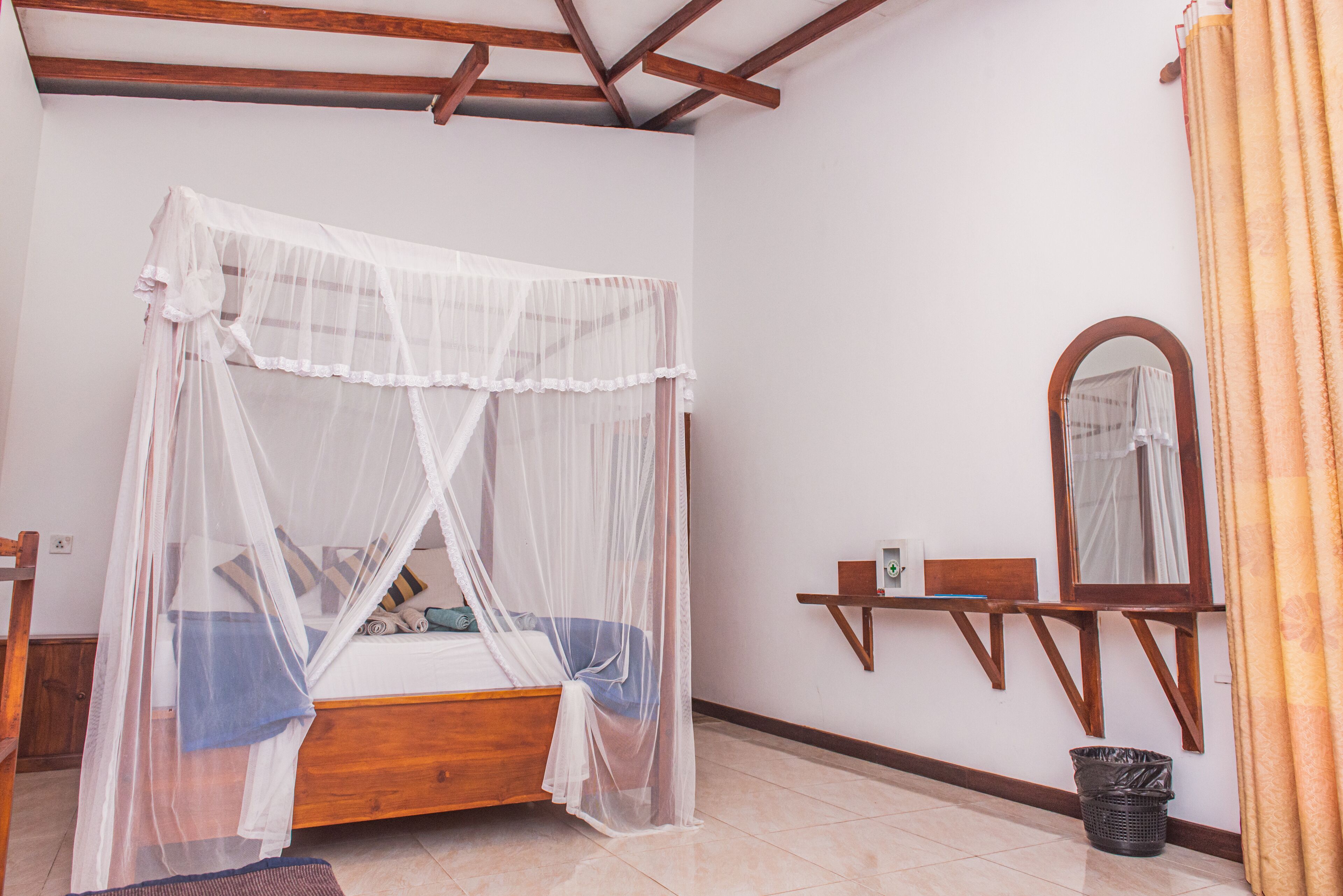 A bedroom with a wooden bed, a canopy, a bedside table, a mirror, and a trash bin.