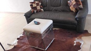 Un salon avec un canapé en cuir, deux coussins décoratifs à motifs, un pouf blanc et un grand tapis en cuir marron et blanc.