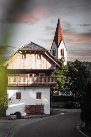 A traditional wooden building with a balcony, a stone wall, and a church with a red roof in the background. - Rousa Little Guesthouse Cosy – Breakfast Included! (Rasun Anterselva)