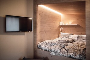 A bedroom with a bed, a flat-screen TV, a shelf with books and a lamp, and a wooden wall.