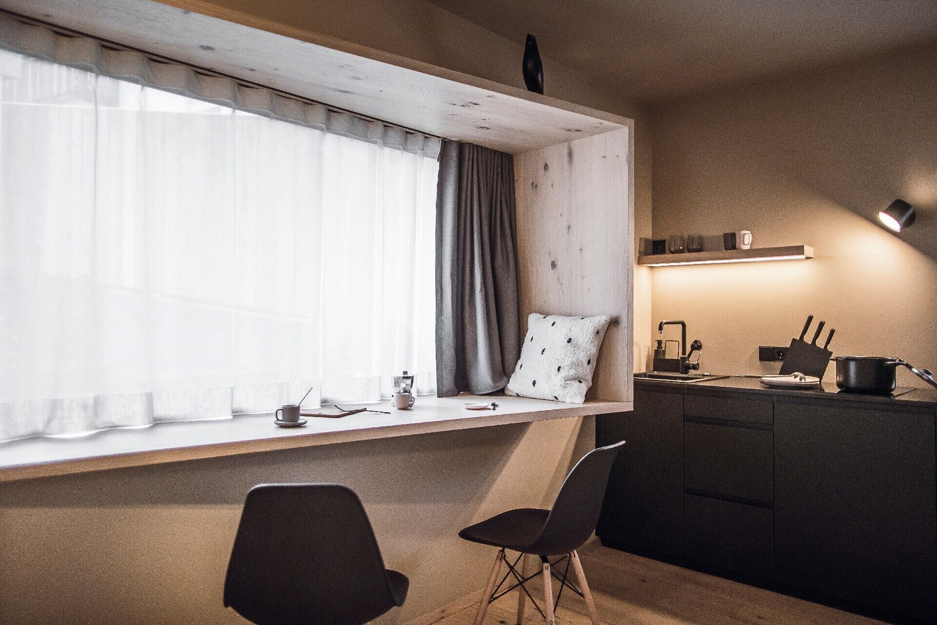 Una cocina compacta con ventana integrada, un pequeño comedor con dos sillas y una encimera con hervidor y utensilios.