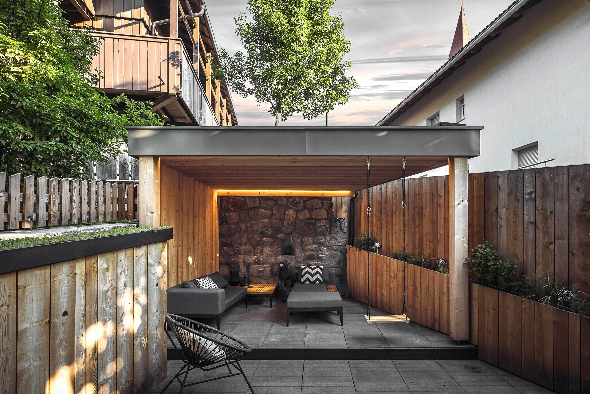 Un espace extérieur abrité, doté d’un mur en pierre, d’une terrasse en bois et d’une chaise en métal noir.