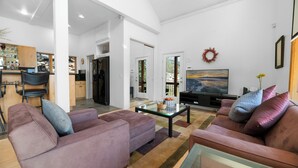 A spacious living room with a large sofa, a coffee table, and a television. There is a kitchen area in the background with a dining table and chairs. - Amazing Grace - Fireplace, Jet Tub, Deck, Lake View (Fawnskin)