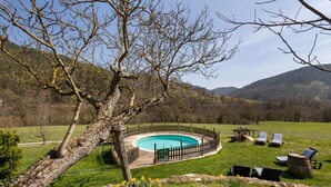 Ein runder Pool mit Holzterrasse und Liegestühlen in einer grünen Umgebung, im Hintergrund Berge.