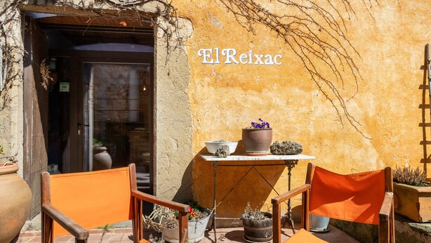 An outdoor seating area with orange chairs, a table with potted plants, and a building entrance with a sign reading 'ElReixac'.