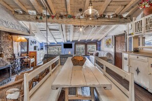 A rustic interior with exposed wooden beams, a central wooden table, wicker furniture, and a television mounted on the wall.