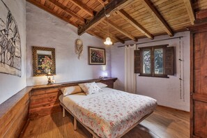 Un dormitorio con una cama de madera, una mesita de noche, una lámpara de pared, un ventanal con contraventanas y techo de madera.