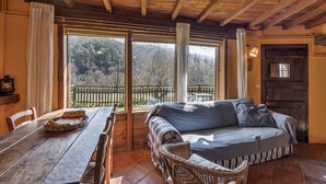 A cozy room with a wooden ceiling, a sofa, a dining table, and large windows offering a view of a wooden deck and trees.