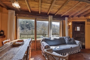A cozy room with a wooden ceiling, a sofa, a dining table, and large windows offering a view of a wooden deck and trees.
