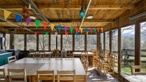 A rustic wooden dining area with large tables and chairs, surrounded by large windows offering a view of the outdoors.