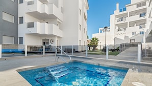 A swimming pool with a metal ladder, surrounded by a tiled deck and white buildings with balconies. - Sungolfbeach El Girasol (orihuela)