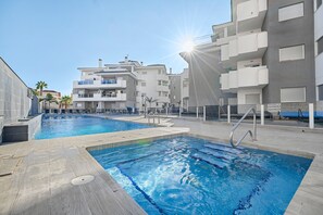 A swimming pool area with two pools, a sunlit sky, and modern buildings in the background. - Sungolfbeach El Girasol (orihuela)