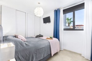 A modern bedroom with a bed, a flat-screen TV, a window with curtains, and a view of buildings outside. - Sungolfbeach El Girasol (orihuela)
