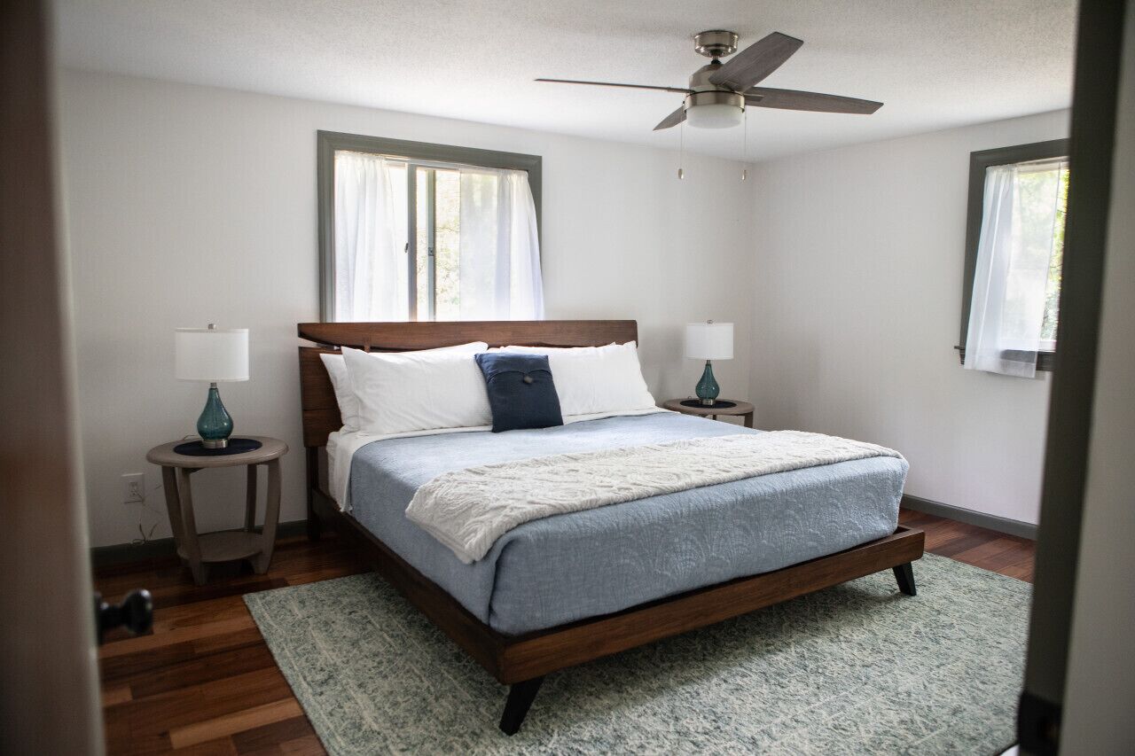 A bedroom with a wooden bed, two bedside tables, a ceiling fan, and windows with curtains.
