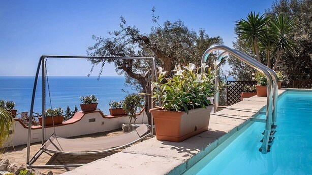 Zona de piscina con sillón colgante, plantas en macetas y vista al mar.