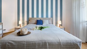 Una cama bien hecha con una colcha gris, un cabecero de madera, dos lámparas de noche y una pared con rayas verticales azules y blancas.