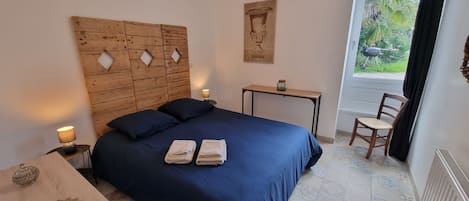 Un dormitorio con cabecera de madera, cama con ropa de cama azul, dos mesitas de noche con lámparas, una mesita pequeña con un tarro y una silla.