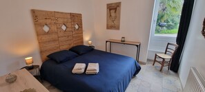 Une chambre à coucher avec une tête de lit en bois, un lit avec une literie bleue, deux tables de chevet avec des lampes, une petite table sur laquelle se trouve un pot, et une chaise.