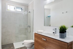 A modern bathroom with a glass shower enclosure, a toilet, a wooden vanity with a mirror, and a small potted plant.