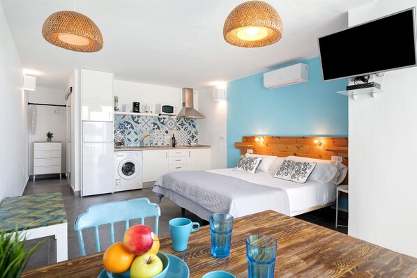 A modern hotel room with a bed, a dining table with fruit, a kitchen area, and a TV mounted on the wall.
