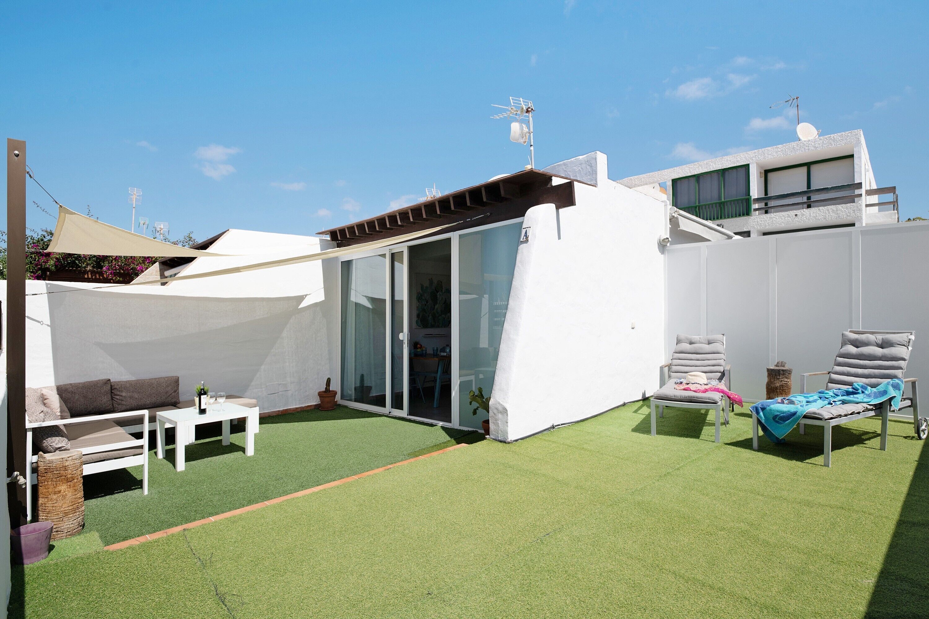 Una terraza en la azotea con césped artificial, un sofá blanco para exteriores, dos sillones de descanso y un edificio blanco con puerta de vidrio.