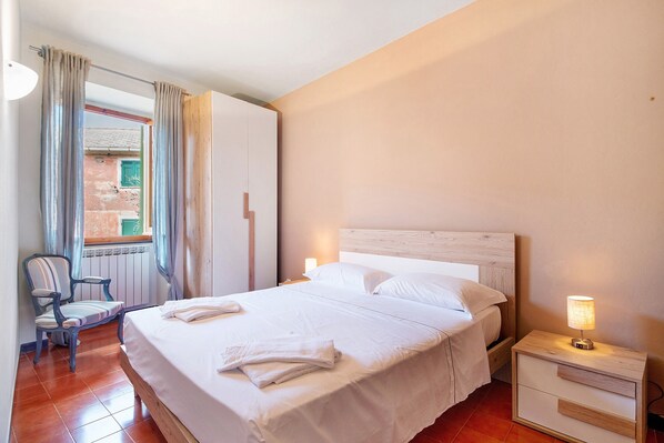 Un lit bien fait, avec du linge de lit blanc, une lampe de chevet, une chaise et une vue sur un espace extérieur à travers une fenêtre ornée de rideaux.
